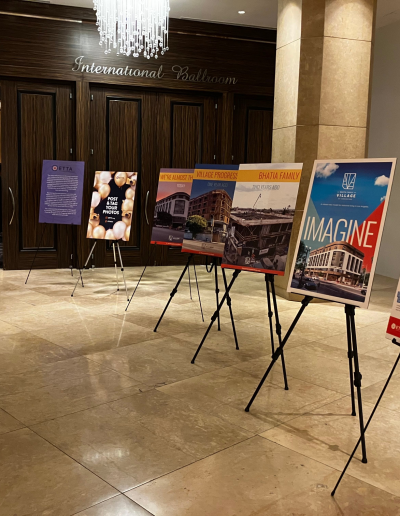 A hallway leading to an open doorway. Inside, people in formal attire are gathered. In the foreground, several colorful event posters on easels line the wall, promoting programs and community initiatives. The floor is polished marble and a chandelier hangs above.